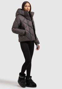 Quiltet grå pufferjakke med høj krave, lynlås lukning og sidelommer. Båret over sorte leggings og sorte støvler med pelsfor.