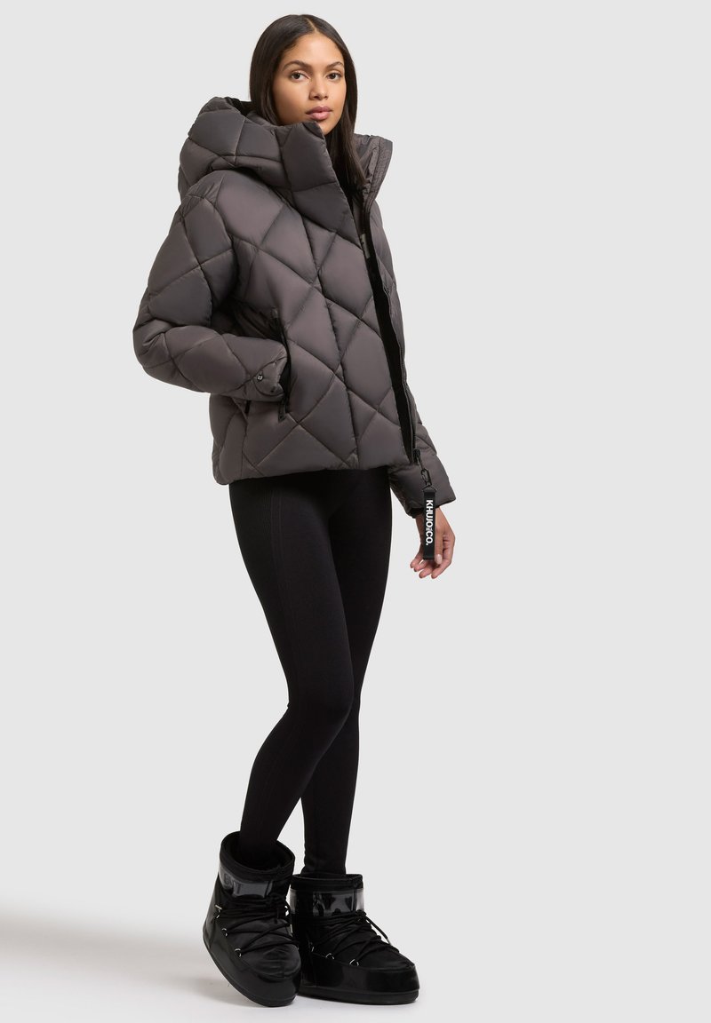 Veste matelassée gris avec col montant, fermeture éclair et poches latérales. Portée par-dessus des leggings noirs et des bottes doublées en fourrure noires.