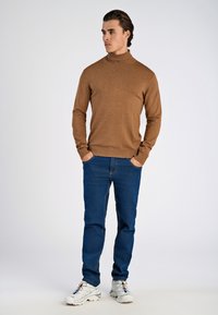 Maglione marrone a collo alto a maglia, jeans blu e scarpe da ginnastica bianche. Il maglione ha un design aderente e polsini a coste; texture liscia.
