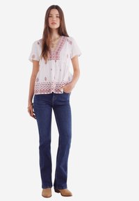 Witte blouse met rode geometrische borduursels, korte mouwen en een V-hals, gecombineerd met donkerblauwe flared jeans en beige enkellaarsjes.