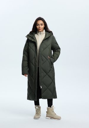 Larga chaqueta de plumas verde oliva con acolchado en forma de diamante, bolsillos en el frente y capucha, superpuesta sobre un suéter crema de cuello alto y pantalones negros.