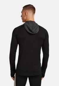 Mand, der vender væk, iført en tætsiddende sort langærmet hættetrøje med hætte i mørkegrå, stående foran en ensfarvet lys baggrund.