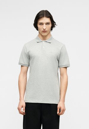 Bărbat tânăr cu păr închis la culoare, purtând un tricou polo gri deschis cu mâneci scurte și pantaloni negri, stând în picioare pe un fond simplu, deschis la culoare.