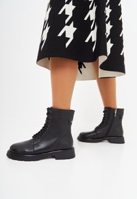 Botas de cuero negras hasta el tobillo con un acabado texturizado, que presentan cordones, cremallera lateral y una suela gruesa. Llevadas con una falda de patrón en contraste.