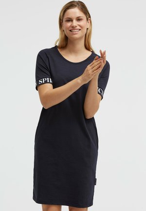 Lächelnde Frau mit blonden Haaren, die ein schwarzes Kleid mit kurzen Ärmeln und weißem Text auf den Ärmeln trägt, steht vor einem schlichten hellen Hintergrund.