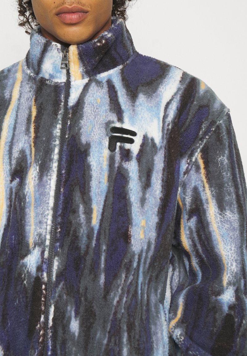 Giacca in pile di blu, viola e giallo, con un colletto alto, cerniera completa e dettaglio del logo nero sul petto. Texture morbida, motivo astratto.