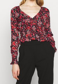 Schwarze Bluse mit floralem Muster in Rot- und Rosatönen, mit V-Ausschnitt, gerafften Details und Rüschensäumen, kombiniert mit schwarzen Hosen.