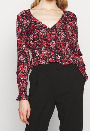 Schwarze Bluse mit floralem Muster in Rot- und Rosatönen, mit V-Ausschnitt, gerafften Details und Rüschensäumen, kombiniert mit schwarzen Hosen.