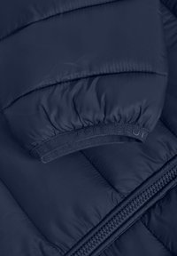 Marinblå puffjacka med quiltat mönster, med ribbat manschett och "POLO CLUB"-logotyp samt dragkedja. Slät, glänsande tygtextur.