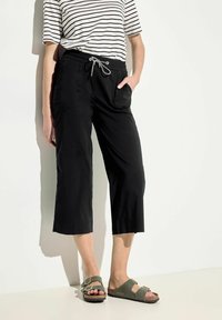 Schwarze, kurz geschnittene Hose mit elastischem Bund und Kordelzug, ausgestattet mit seitlichen Taschen und einem lockeren Schnitt. Kombiniert mit olivgrünen Sandalen.