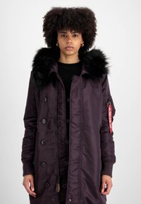 Ljubičasti podstavljeni parka s kapuljačom od umjetnog krzna, prednjim dugmadi i rebrastim manžetama. Ima crvenu oznaku na rukavu i zatvaranje na zatvarač.