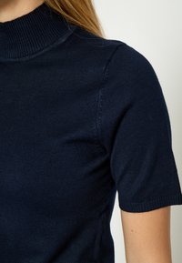 Žena odjevena u tamnoplavi prsluk s rebrastim uzorkom i visokim okovratnikom kratkih rukava, prikazuje gornji torzo i detalje ramena.