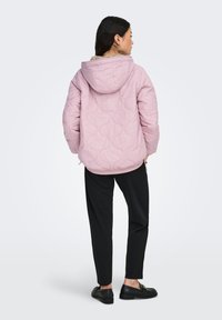 Vaaleanpunainen tikattu takki hupulla, jossa on teksturoitu kuvio. Malli pukeutunut mustiin housuihin ja mustiin kenkiin, selin kameraan.