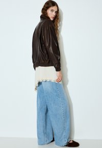 Braune Lederjacke mit tailliertem Schnitt, kombiniert mit einem cremefarbenen, mit Spitze verzierten Top und weit geschnittenen hellblauen Jeans. Braune, flauschige Slipper.