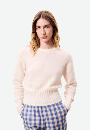 Femme aux cheveux blonds ondulés mi-longs, portant un pull crème et un pantalon à carreaux bleu et blanc, mains dans les poches, fond neutre.