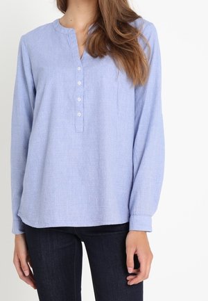 Kobieta ubrana w jasnoniebieską, długorękawową, zapinaną na guziki bluzkę z okrągłym dekoltem oraz ciemne spodnie na jednolitym białym tle.