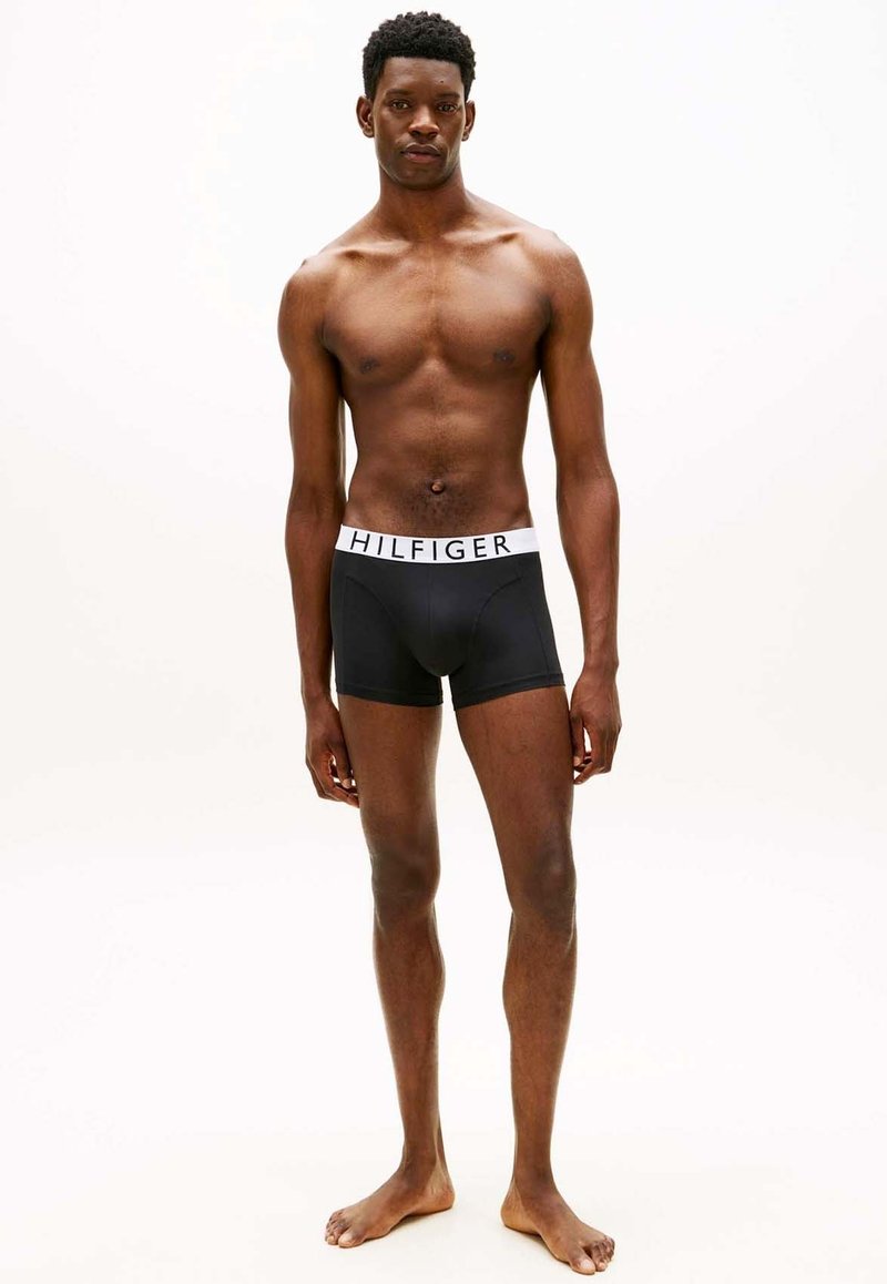 Modèle masculin noir mince, debout pieds nus, portant un boxer noir Tommy Hilfiger avec une ceinture blanche marquée, sur fond blanc uni.