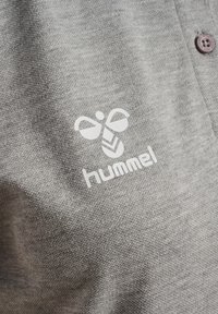 Grå tekstil polo-shirt med hvidt Hummel-logo og knapdetalje. Det teksturerede stof viser et subtilt meleret mønster.