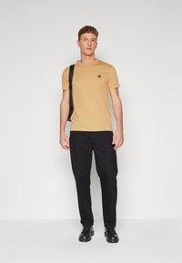 Kortærmet t-shirt med runde hals i lys brun med et logo på brystet, parret med sorte bukser og sorte sko. Simpelt design.
