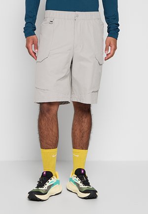 Homme portant un short cargo beige, des chaussettes jaunes Nike et des chaussures de sport multicolores, debout devant un fond uni.