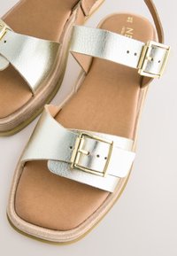 Sølvfarvede metalliske sandaler med en tekstureret finish, der har to brede remme og en guldbeklædt spænde detalje. Polstret fodseng og en kraftig sål.