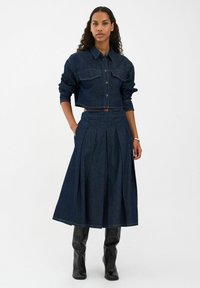 Jeans jakna s dva džepa na prsima, uparena s suknjom s visokim strukom i naborima od tamnoplave teksas tkanine. Model nosi crne čizme do koljena.