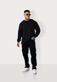 Schwarzes Langarm-T-Shirt mit Rundhals, kombiniert mit schwarzen Cargo-Hosen und weißen Sneakers. Enthält Tätowierungsdetails an den Unterarmen.