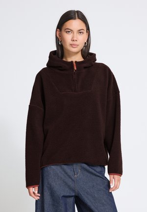 Jeune femme aux cheveux lisses et foncés porte un sweat à capuche polaire noir zippé et un jean bleu large, debout devant un fond clair uni.