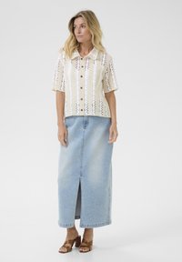 White crochet-style short-sleeve shirt with brown buttons, paired with a light blue denim maxi skirt featuring a front slit. Brown block-heeled sandals.
