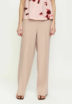 Naine, kes kannab beeže laia säärega pükse, roosat ülaosa punaste lillemustritega ja pruune ruudukujulise varbaga sandaale, seistes valge tausta ees.