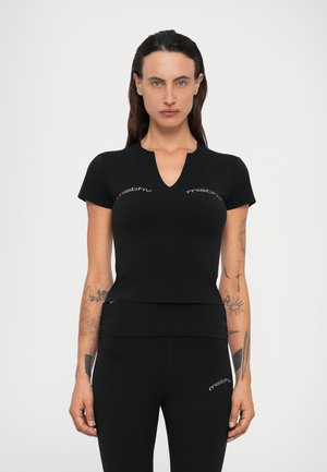 Moteris su ilgais plaukais, vilkinti juodą prigludusį V-formos iškirptės trumpomis rankovėmis marškinėlį ir juodas leggins, abu su "misbhv" logotipu, stovi prieš paprastą foną.