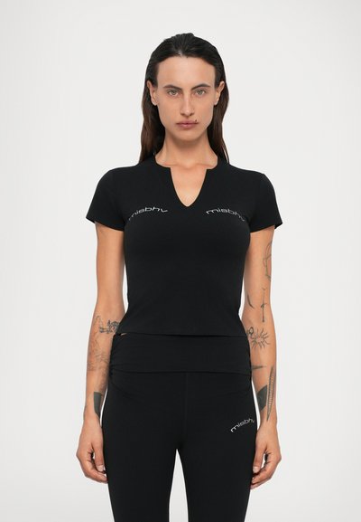 Femme aux longs cheveux portant un haut à col en V noir ajusté à manches courtes et un legging noir, tous deux avec le logo "misbhv", se tenant devant un fond uni.
