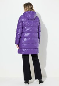 Chaqueta acolchada morada con capucha, con un acabado brillante, acolchado horizontal y mangas largas. Pantalones negros y tacones completan el conjunto.