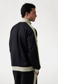 Veste de sport noire avec un accent à rayures gris clair. Elle présente un col et des poignets côtelés, une texture de tissu lisse et une coupe décontractée.