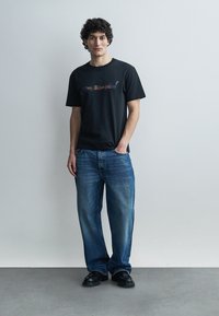 Νέος άνδρας με σγουρά μαλλιά, φοράει μαύρο μπλουζάκι με σχέδιο, μπλε φαρδιά τζιν και μαύρα παπούτσια, στέκεται μπροστά σε απλό ανοιχτό γκρι φόντο.