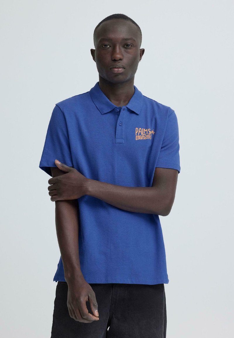 Jeune homme debout avec une expression neutre, portant un polo bleu avec le logo "Palms Island", la main reposant sur le bras opposé.