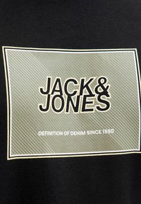 Sort T-shirt med et rektangulært logo i olivengrøn, der viser "JACK & JONES" i fed sort skrift. Teksten inkluderer "DEFINITION OF DENIM SINCE 1990."