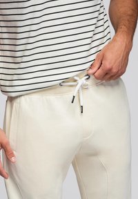 Světlé krémové tepláky s bílou šňůrkou a černými kovovými detaily. Tkanina vypadá měkce, má boční kapsy a tapered leg design.