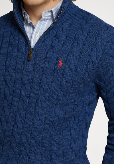 Μπλε ναυτικό πουλόβερ με πλεκτό καλώδιο και γιακά με φερμουάρ στο ένα τέταρτο, με κόκκινο λογότυπο στο στήθος και υφασμένο μοτίβο σε όλο το ύφασμα.