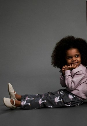 Een jong kind met krullend haar zit op de vloer, draagt een roze sweatshirt, donkere broek met witte vogelprint en lichtgekleurde schoenen, en glimlacht verlegen.