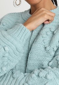 Närbild av en person som bär en mjuk, ljusblå stickad tröja med texturerade mönster, där deras hand vilar nära kragen och en stor örhänge.