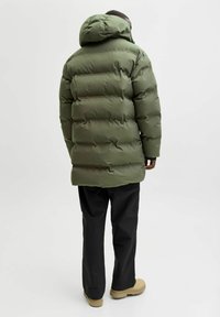 Person som står vänd bortåt, kl�ädd i en olivgrön vadderad jacka med huva, svarta byxor och beige stövlar, mot en enfärgad bakgrund.