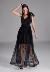 Robe de soirée noire avec un corsage ajusté et une superposition transparente. Présente un décolleté en V profond, un détail en nœud sur une épaule, et un tissu délicatement imprimé.