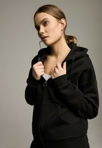 Czarna bluza z kapturem na zamek, wykonana z miękkiego materiału, z przednią kieszenią, sznurkiem ściągającym i ściągaczami przy mankietach. Model trzyma zamek.