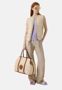 Padded creamfarbener Weste, helllila Oberteil, beiges weitgeschnittenes Hosen, trägt eine hellbraune geflochtene Tasche mit braunen Akzenten und einem runden Logo. Winkelige Sonnenbrille.