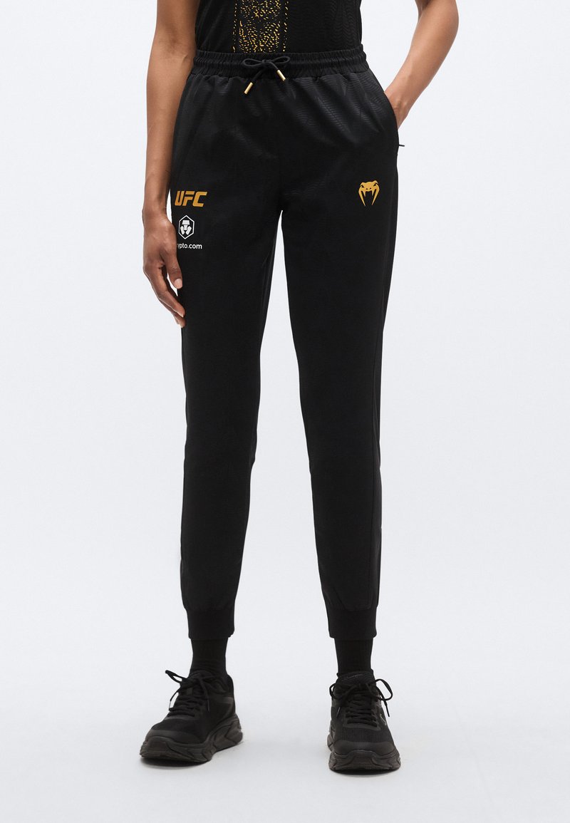 Pantalon de sport noir à coupe fuselée, avec taille à cordon, poignets côtelés et logos dorés sur la cuisse gauche et le côté.