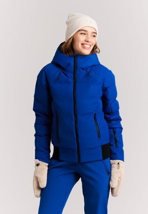 Jonge vrouw lacht, draagt een blauwe winterjas en broek, een beige gebreide muts en beige fleece wanten tegen een egale achtergrond.