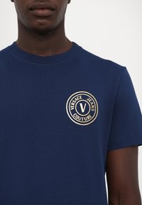 Tums zilas T-kreklas, izgatavots no kokvilnas, ar apaļu izgriezumu. Uz krūtīm ir zelta apļa logo ar tekstu "VERSACE JEANS COUTURE".