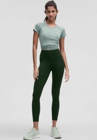 Gaiši zaļš pieguļošs T-krekls, apvienots ar augsto vidukli tumši zaļām legingām un gaiši zaļiem sporta apaviem. Gluda tekstūra, minimāls dizains.