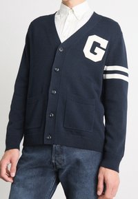 Navyblå kardiagan med V-hals, knappar framtill, två fickor och vit "G"-patch med randiga detaljer på ärmarna. Stickad textur, avslappnad design.